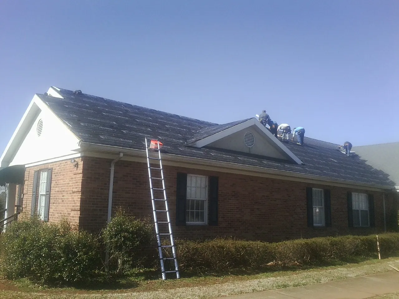 Crew tearing shingles off a brick ranch roof with a ladder set — Emergency Roof Repair work by our crew in Sangerfield