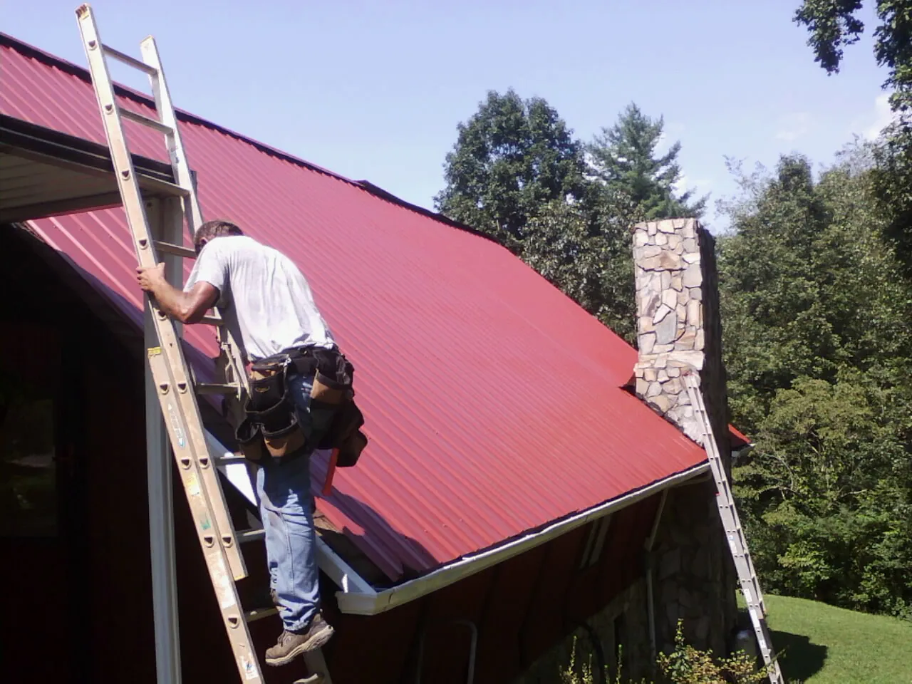 Roofer on a ladder beside a red metal roof and stone chimney — Soffit & Fascia Repair by Roofing Brothers in Sangerfield